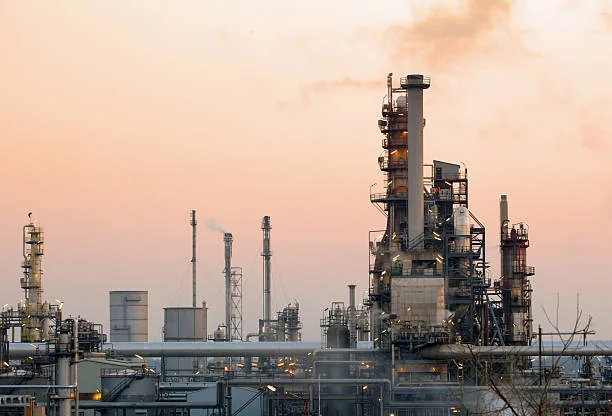 A tall industrial column rising above refinery pipework beneath a hazy sky